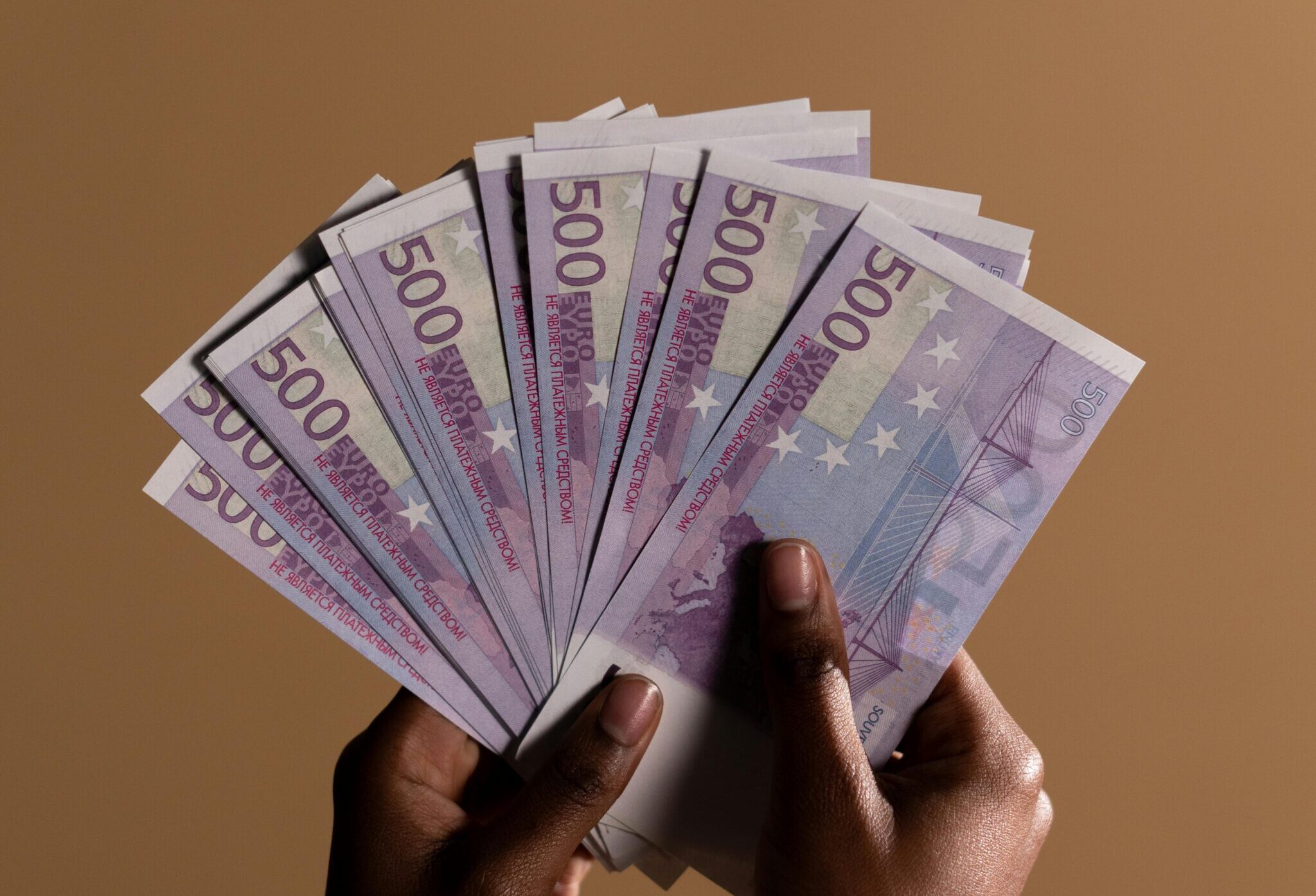 Close-up of hands holding multiple 500 euro banknotes against a beige backdrop.